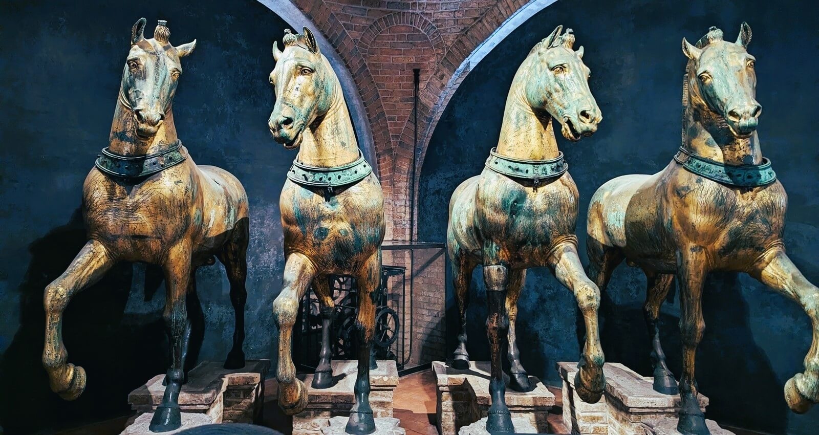 Four metal horses crafted in great detail, they are lined up abreast and appear to be trotting. They look aged with hues of brown and green. 